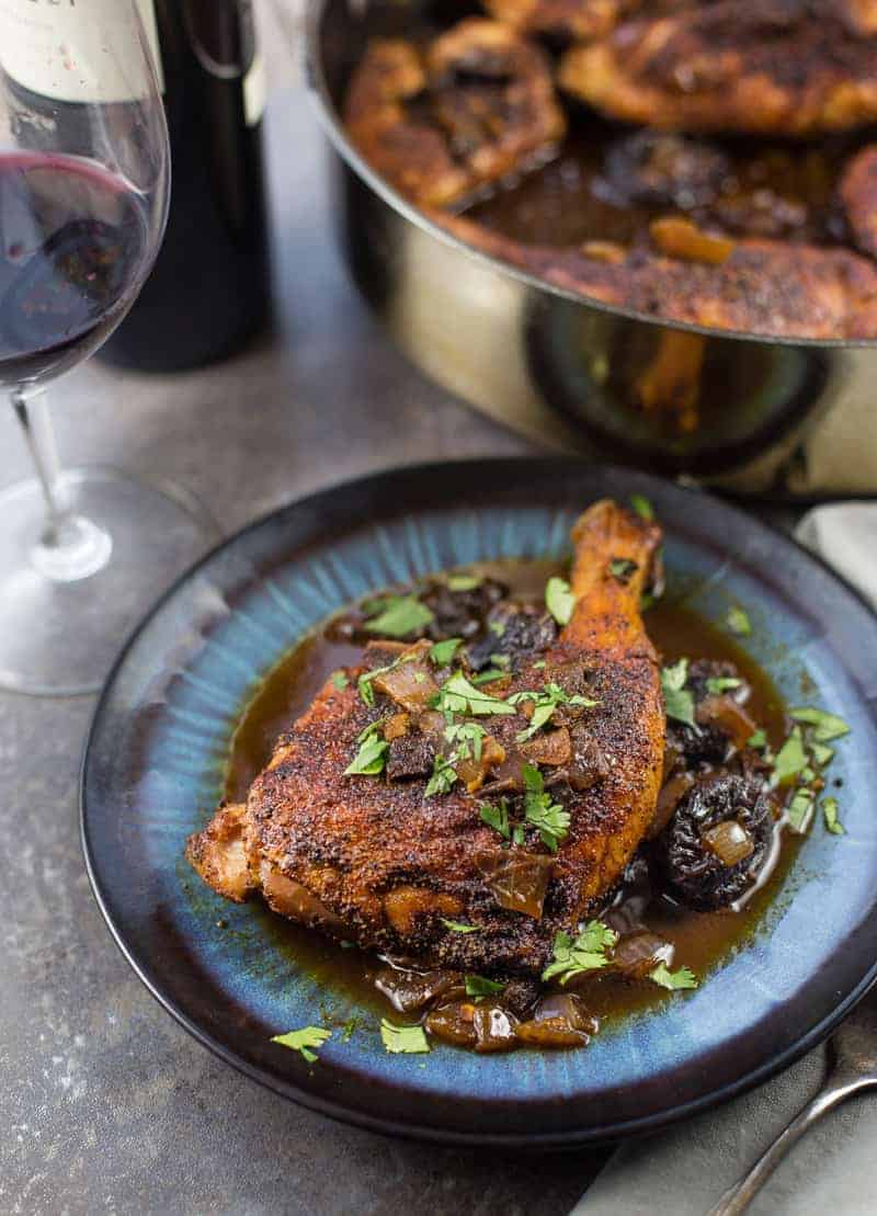 Plated dish with grilled chicken with a wine, ancho chili, and prune sauce with a wine glass on the side.