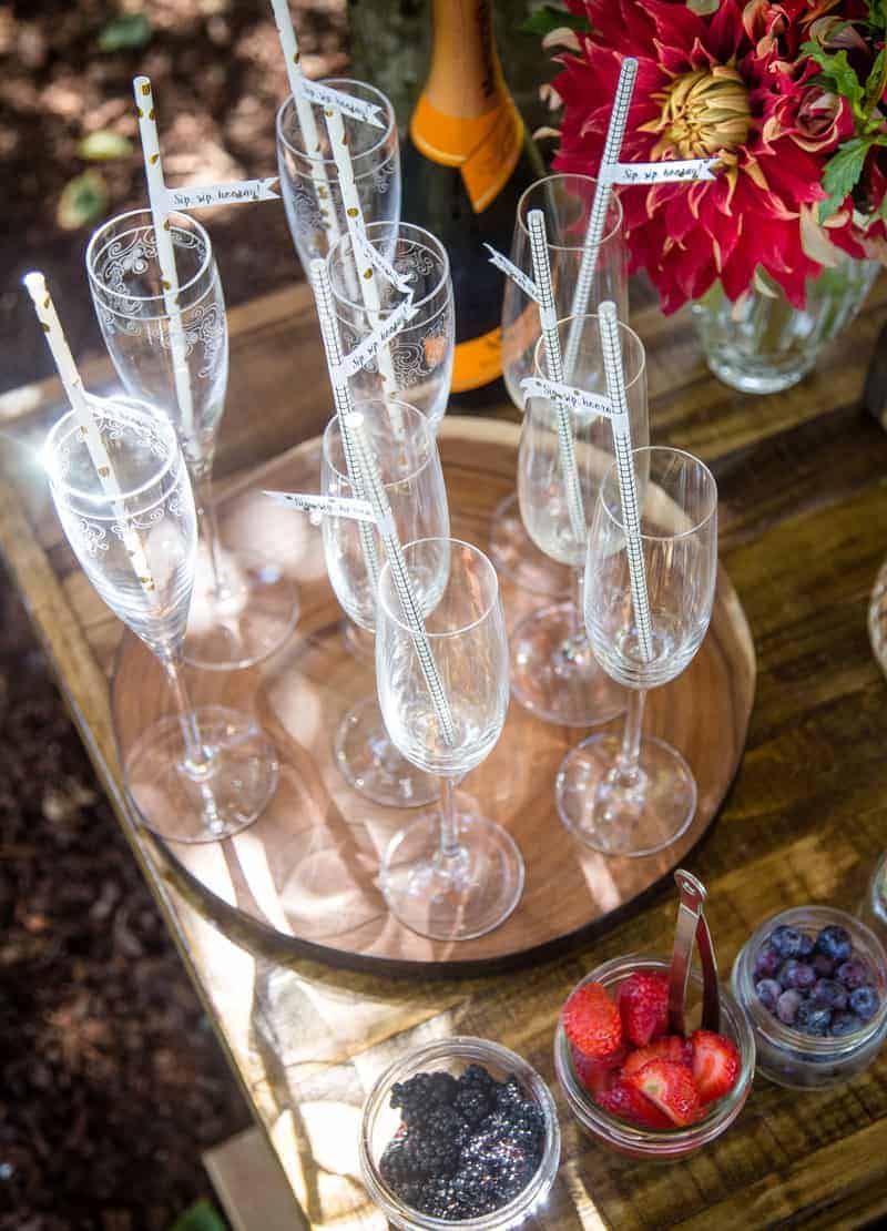 Champagne flutes with straws on a wooden tray with a bottle of prosecco, fruit garnishes in bowls, and a vase of flowers