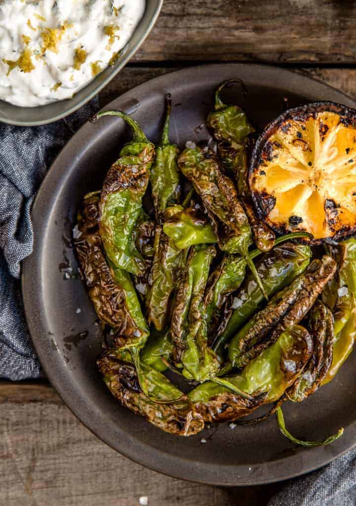 Grilled Shishito Peppers on a plate with a grilled lemon half and lemon yogurt dipping sauce