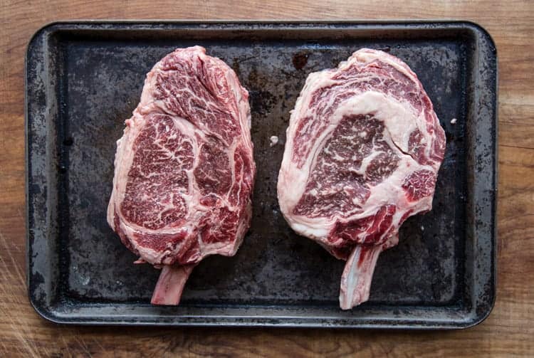 photo of two raw cowboy ribeye steaks on a sheet pan