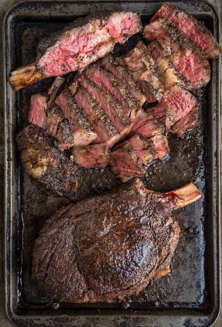 Grilled cowboy ribeye steaks on a sheet pan, one is whole and the other is cut into strips showing the right way to cut a large ribeye