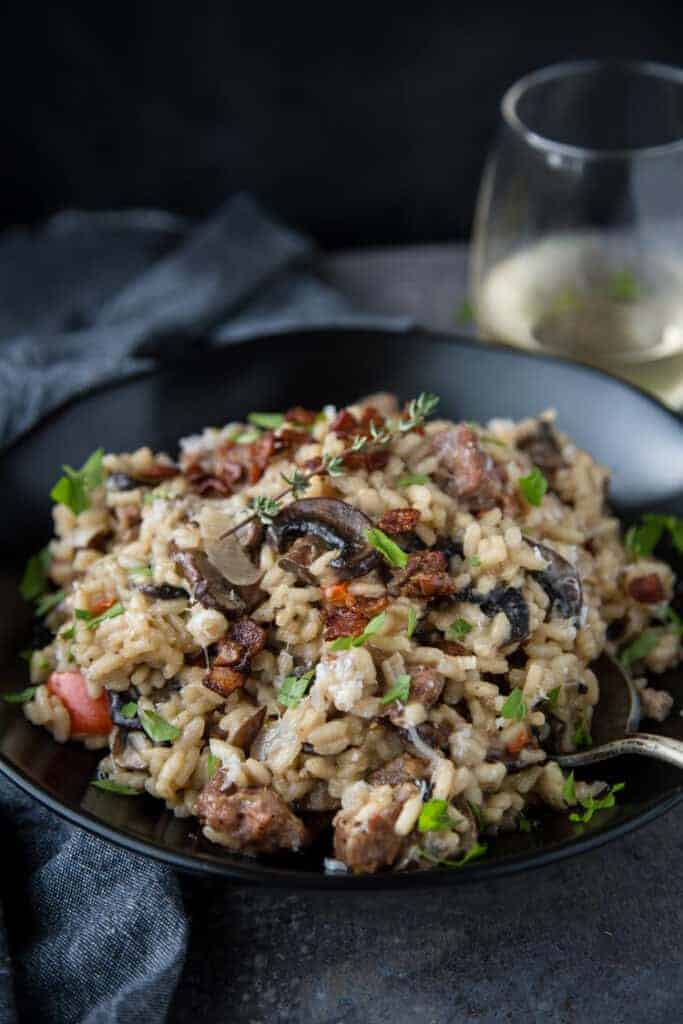 A bowl of homemade risotto with smoked sausage, mushrooms, and bacon, paired with a glass of wine.