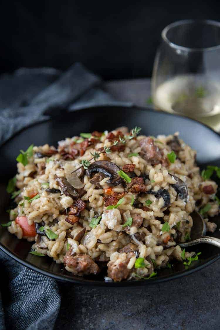A bowl of homemade risotto with smoked sausage, mushrooms, and bacon, paired with a glass of wine. 