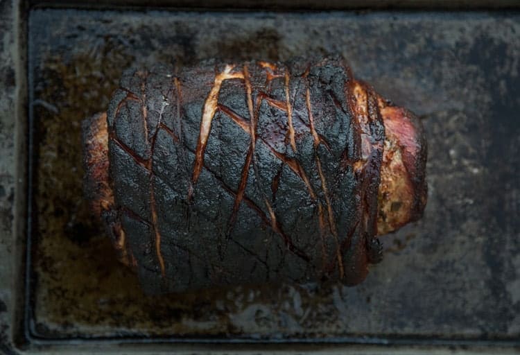 Grilled Porchetta roast resting on a baking sheet. The bark is cut in a diamond pattern