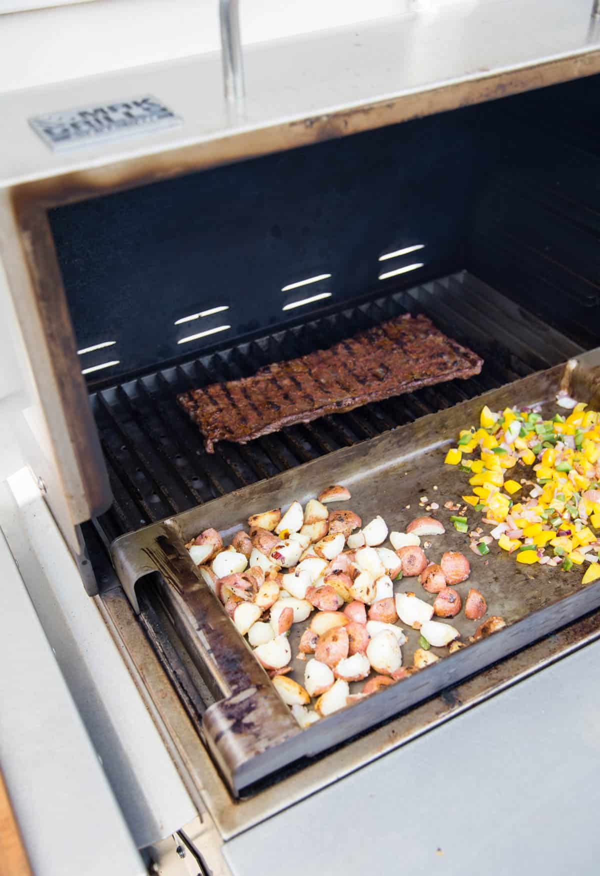 A grill with a skirt steak cooking in the back and potatoes and vegetables cook on a hot griddle in the front