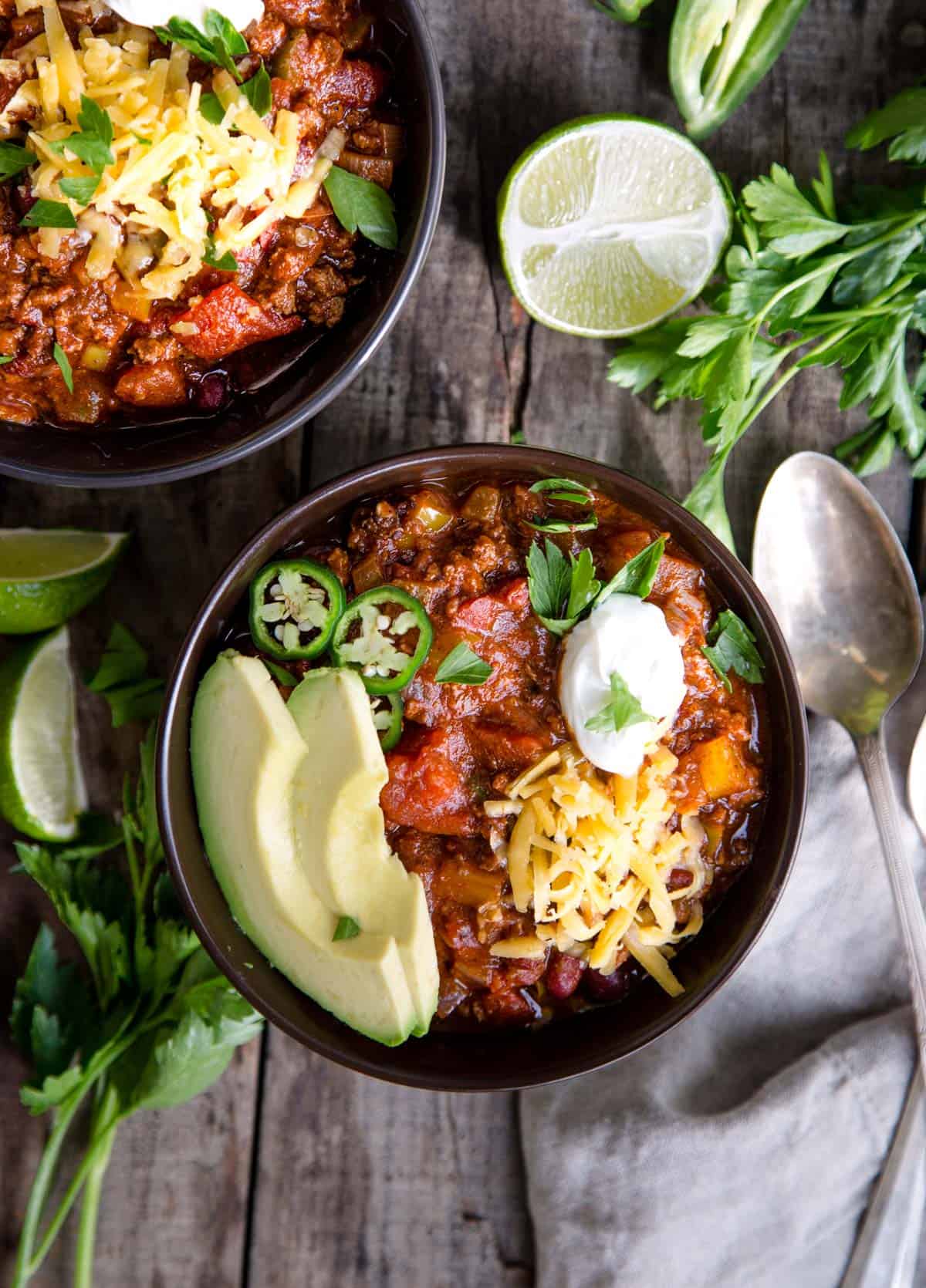 Two bowls of beef chili topped with avocado, jalapeño, cheese, and sour cream. 