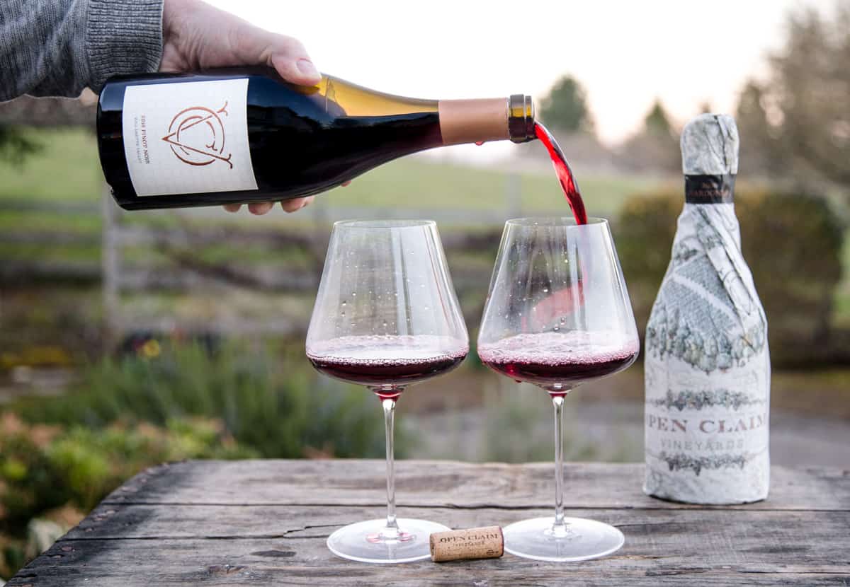 Pouring a bottle of Open Claim Pinot Noir into two wine glasses on a wooden board