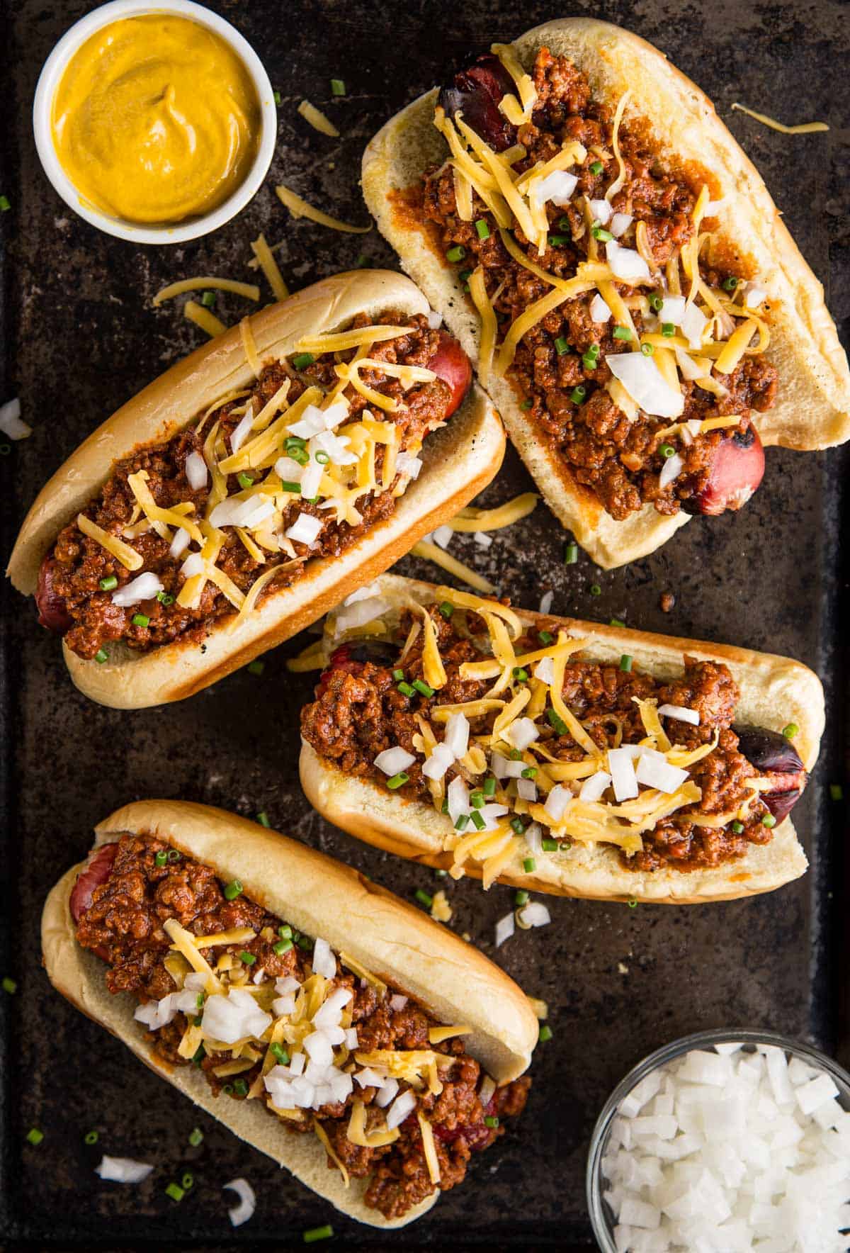 4 Chili Dogs on a platter with a side of yellow mustard and chopped onions