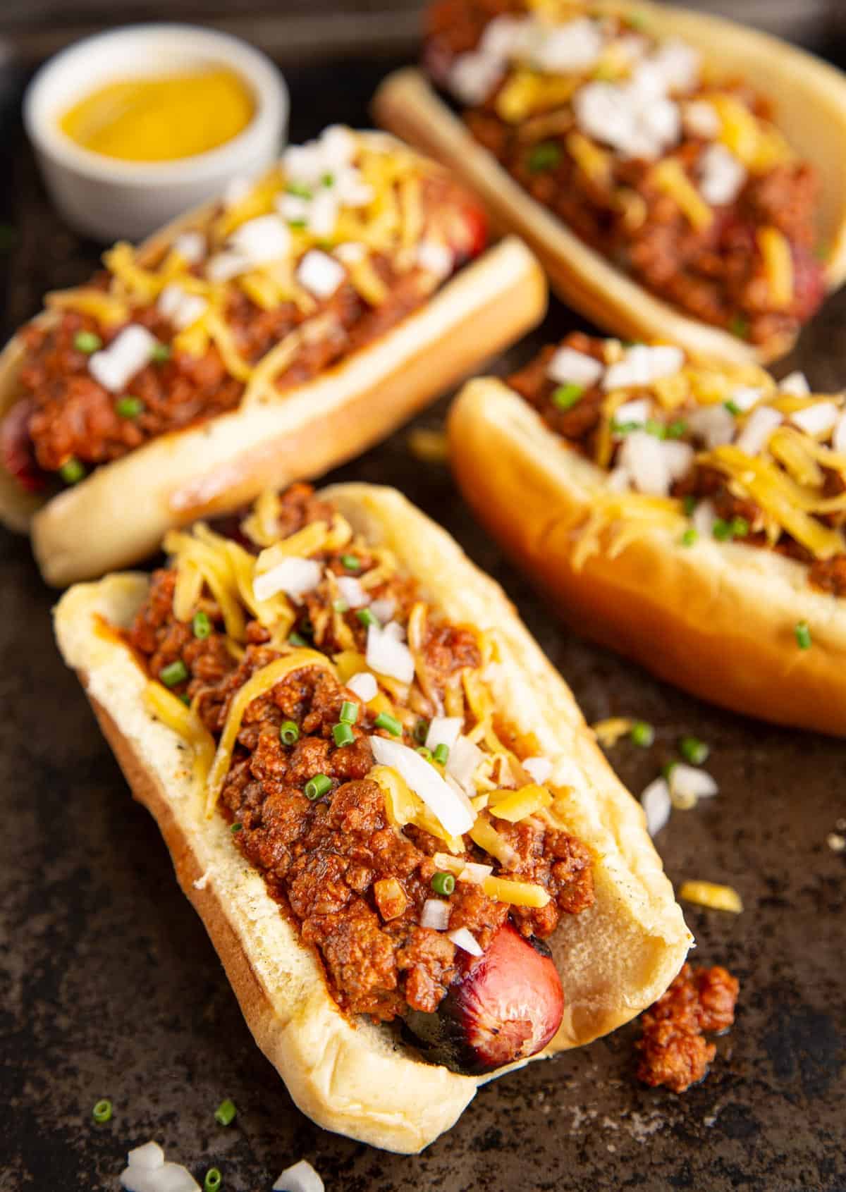 A side angle of chili dogs on a sheet pan topped with shredded cheese and chopped onions.
