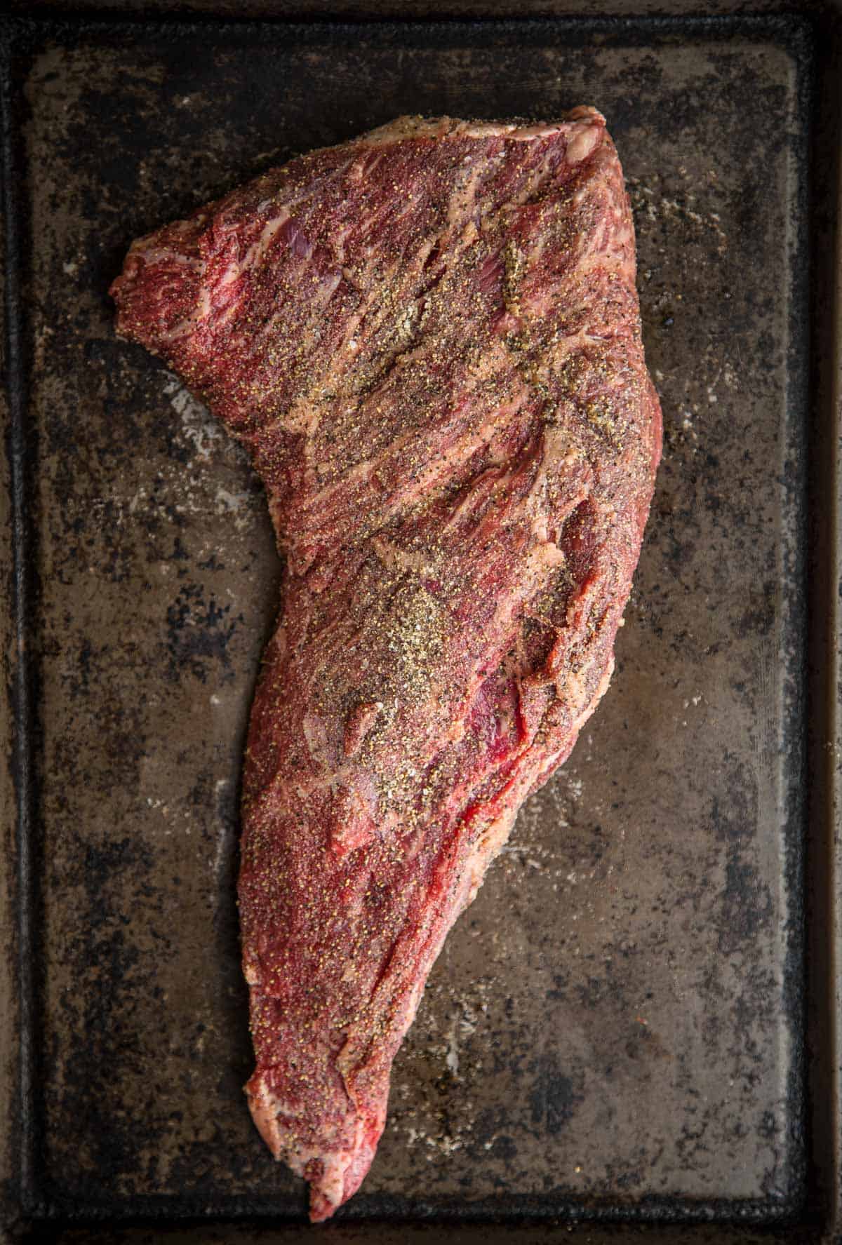 A raw Tri Tip roast on a platter seasoned with salt, pepper, and garlic