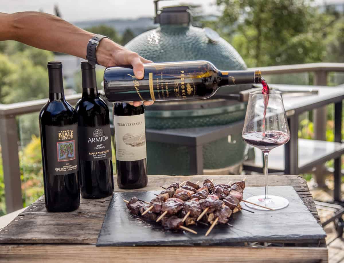 A bottle of Zinfandel being poured into a glass near a plate full of grilled beef skewers.