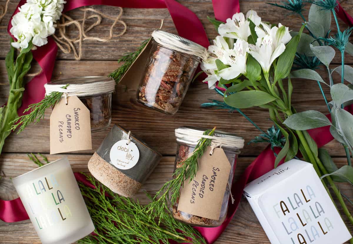 Jars of candied pecans, flowers, and candles, all arranged on a wood board as holiday gift ideas.