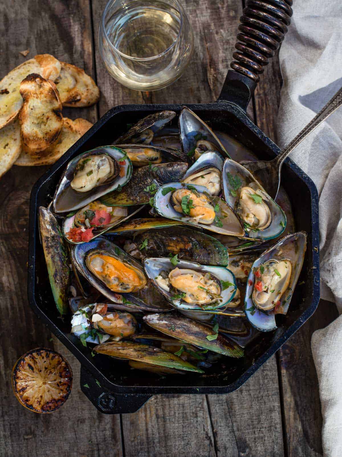 Grilled mussels in a cast iron pan on a wood board with a glass of wine and a grilled lemon halves.