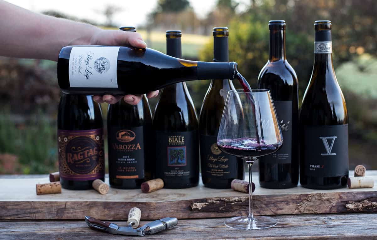 6 bottles of Petit Sirah wine in a lineup, with a bottle being poured into a glass