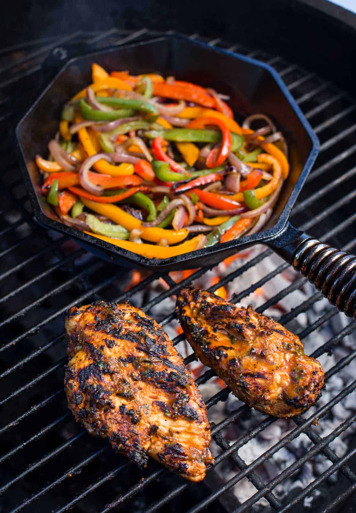 Chicken on the grill alongside peppers and onions cooking in a cast iron pan on the grill.
