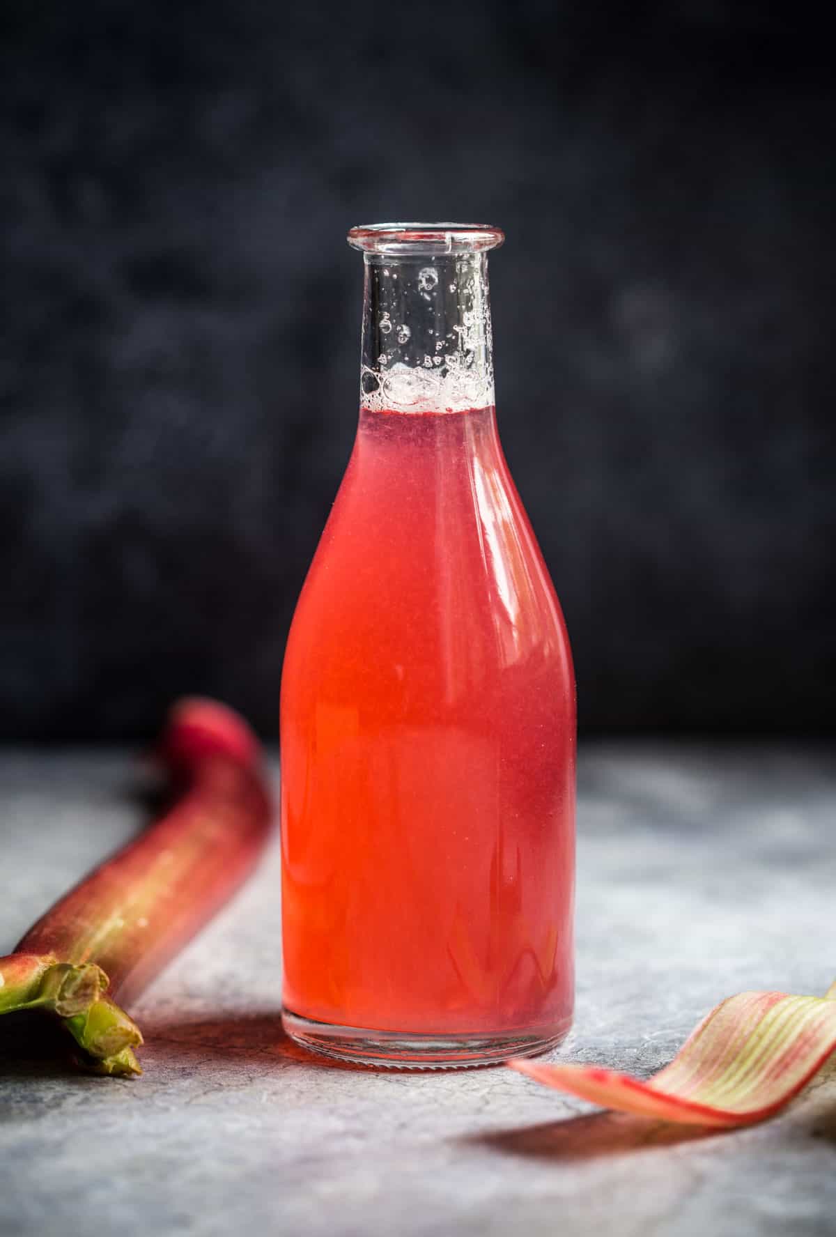 A bottle filled with Rhubarb simple syrup on a table surrounded by rhubarb.