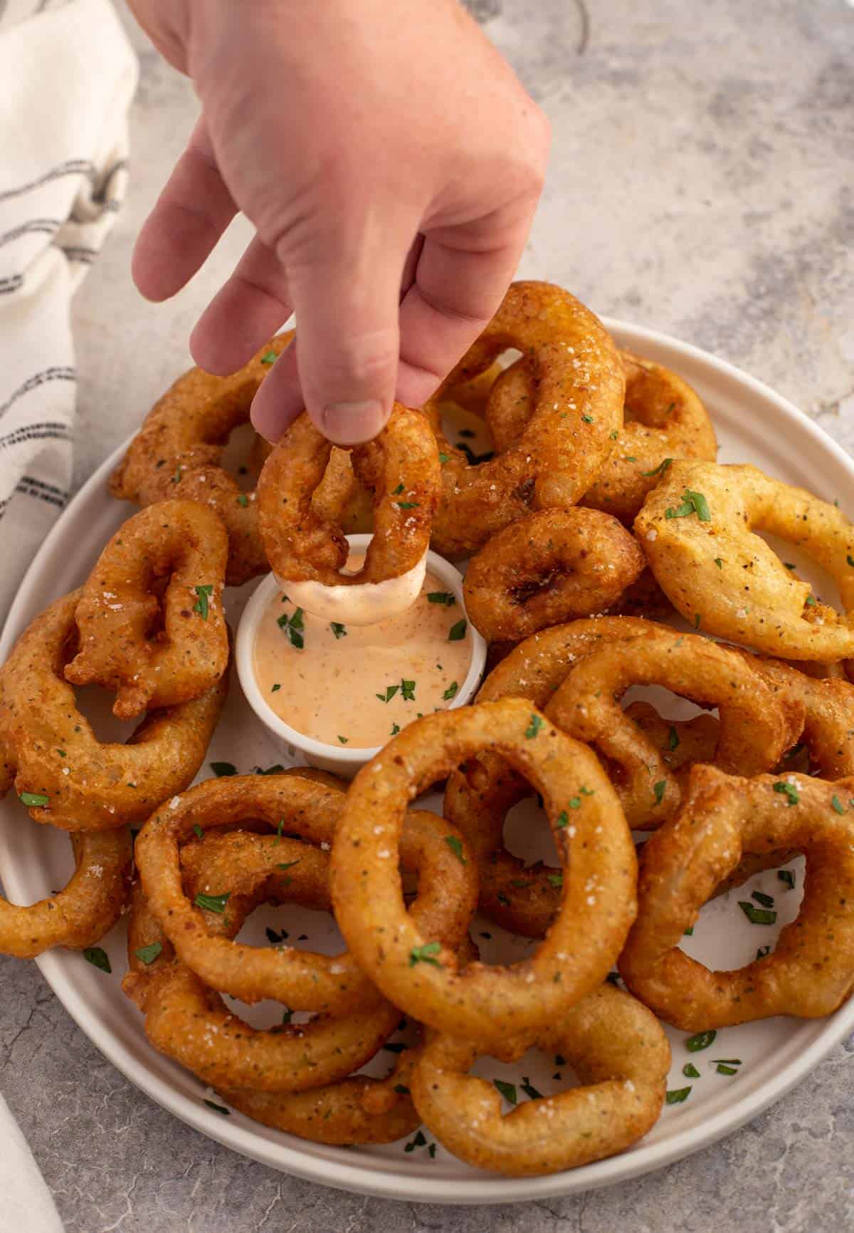 A platter of smoked onion rings and a hand dipping an onion ring into chipotle dipping sauce 