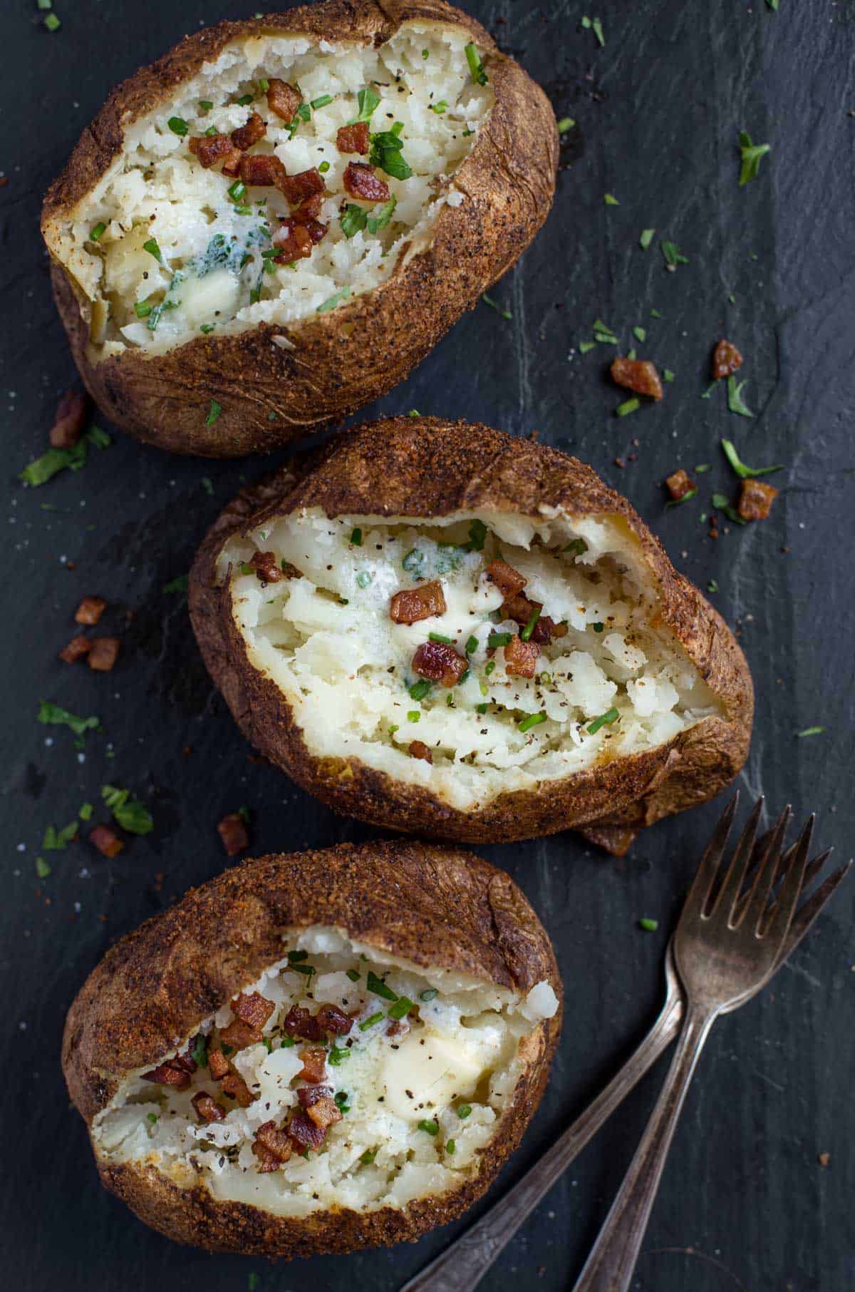 three perfectly smoked baked potatoes on a platter topped with butter, bacon, and chives
