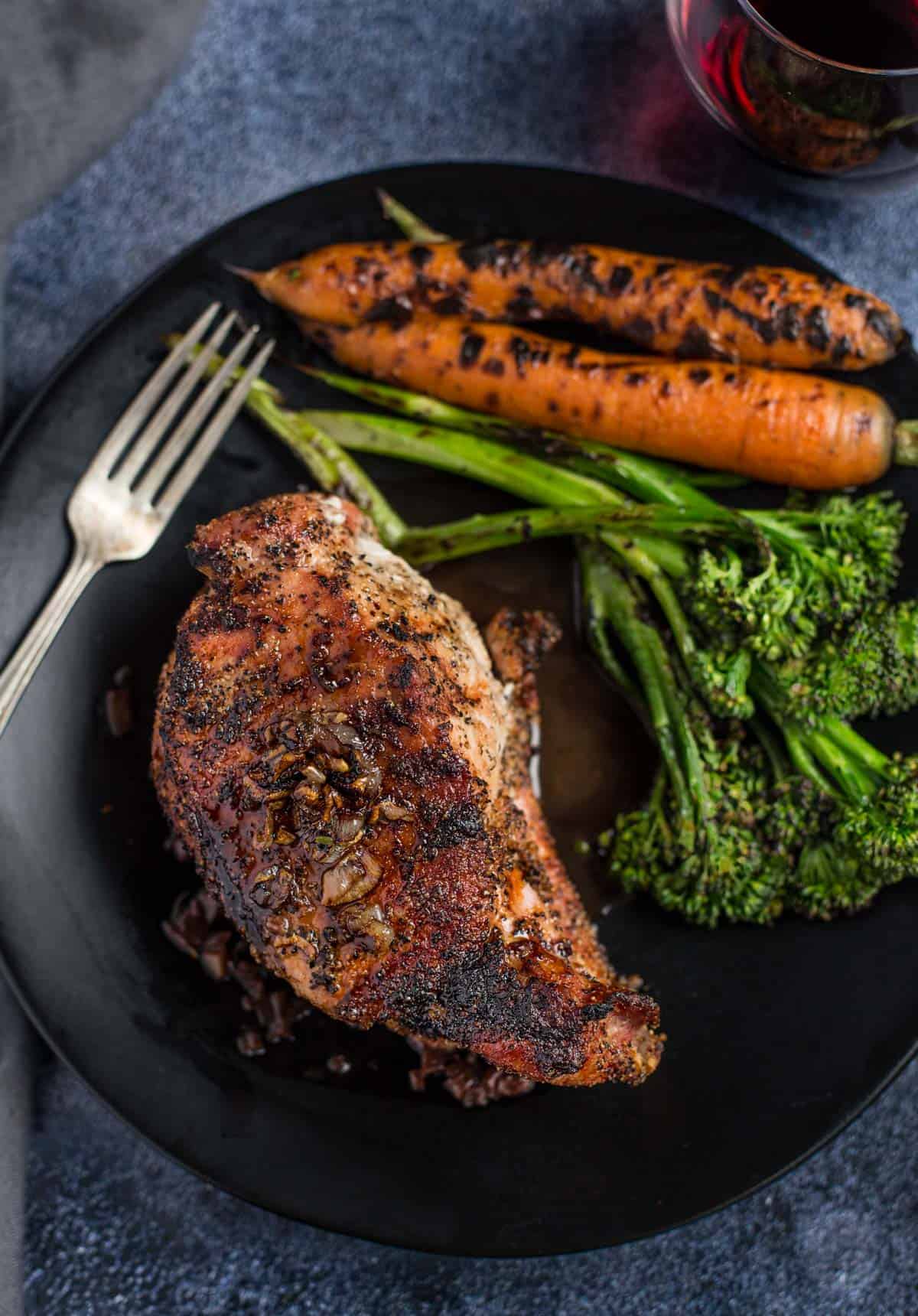 A Grilled Pork Chop drizzled with a red wine pan sauce on a black plate with vegetables on the side