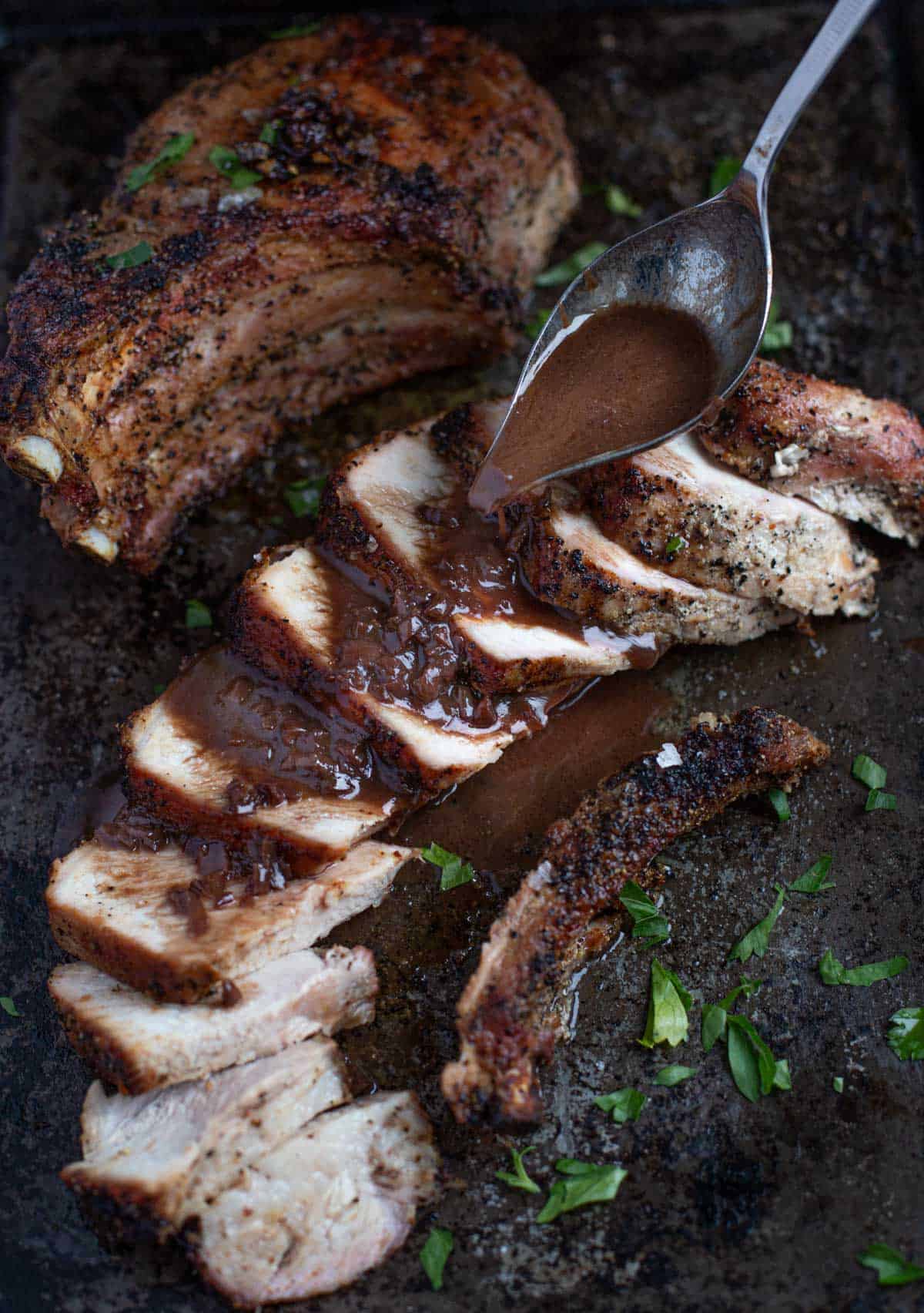 A grilled pork chop on a platter with slices of meat and a spoon pouring red wine pan sauce over the top. 