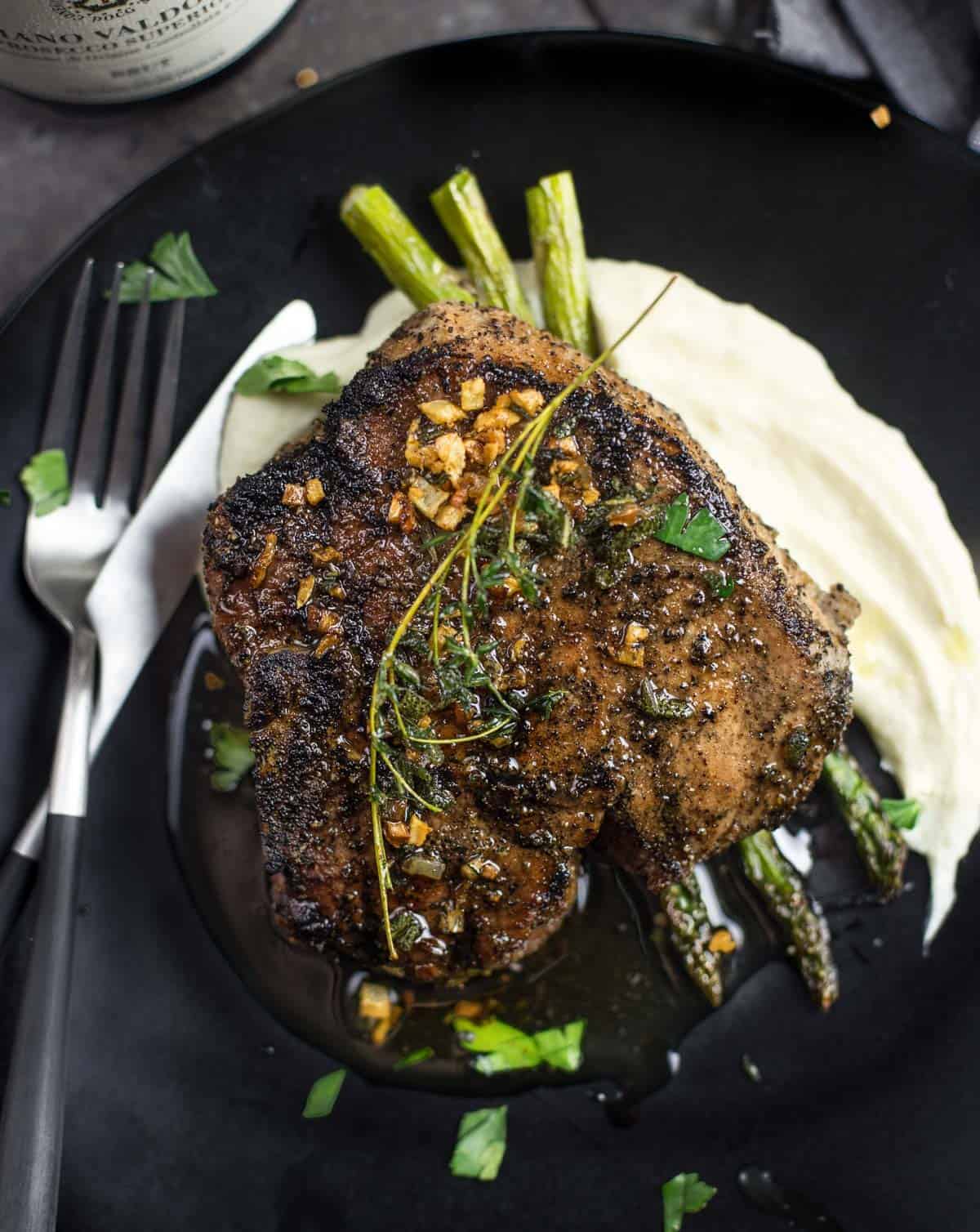 A grilled pork chop topped with a wine brown butter sauce on a plate with a bottle of Prosecco wine