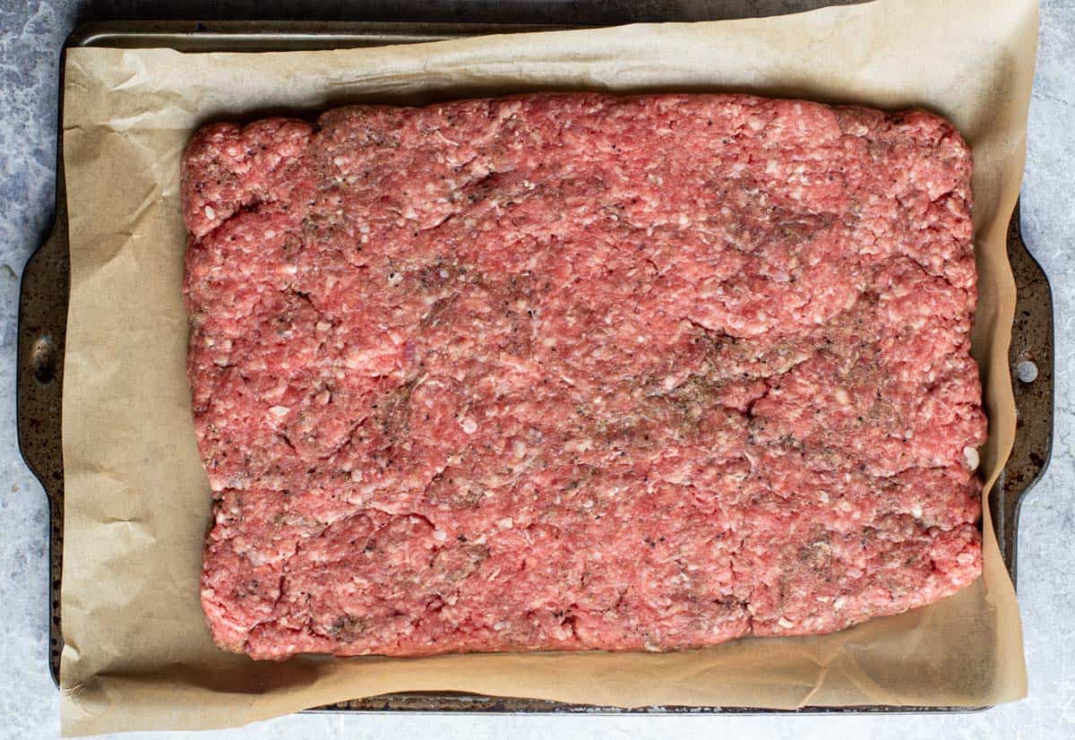 A rectangle of ground beef in preparation for sliders on a sheet tray lined with parchment paper.