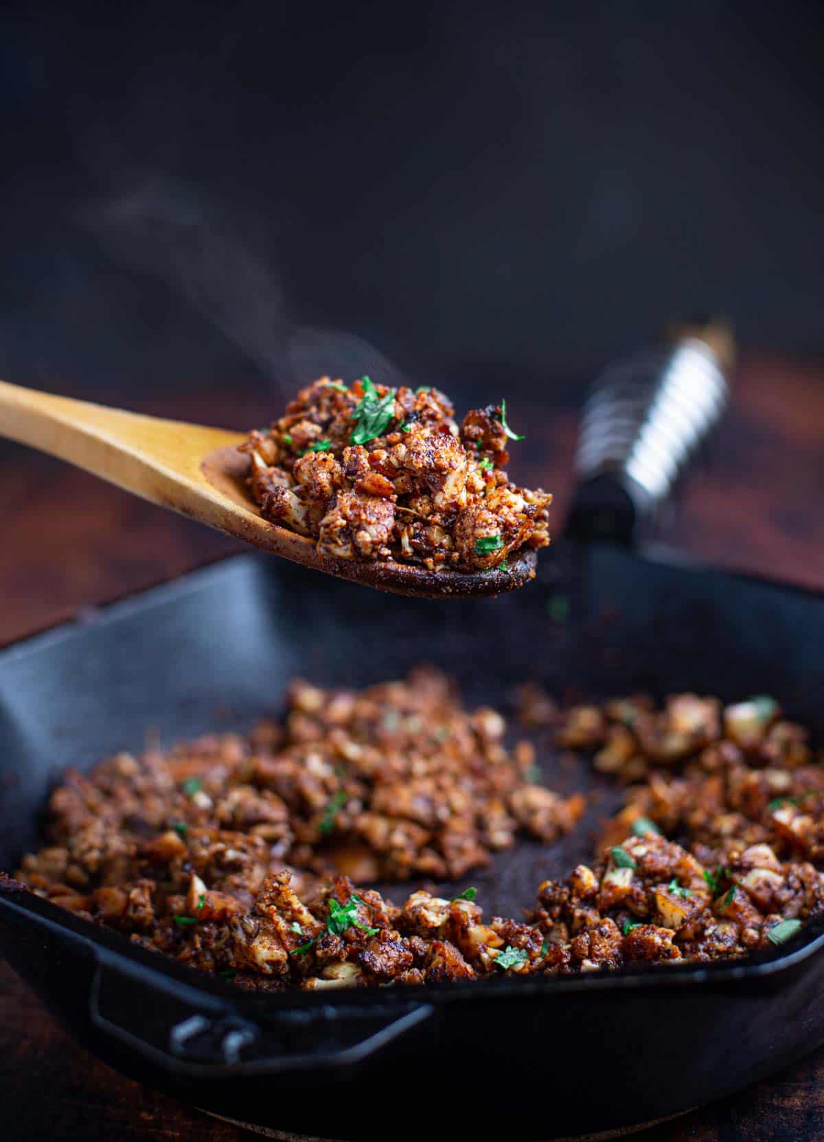 A wooden spoonful of Cauliflower Chorizo topped with parsley taken from a cast iron pan.