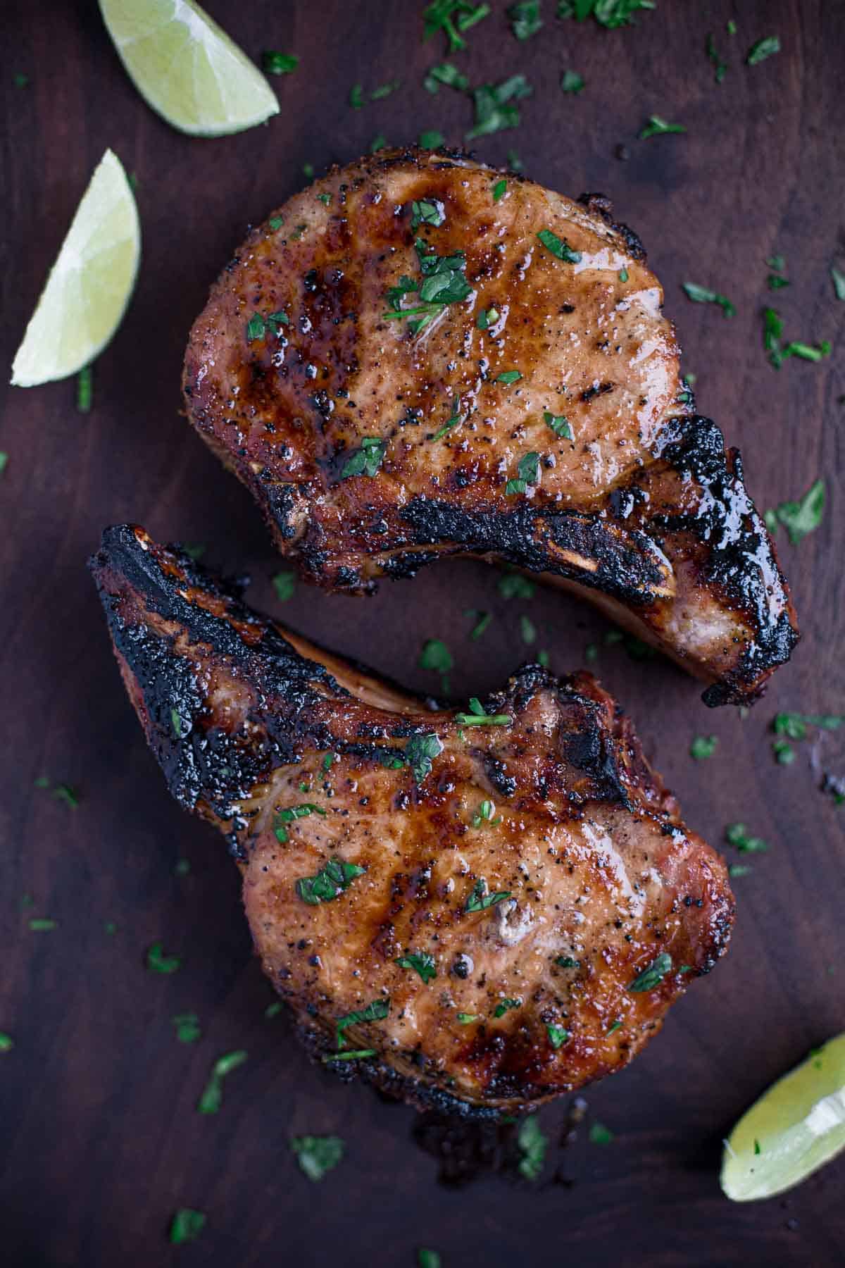 Two grilled pork chops on a wooden cutting board with a wedge of lime.