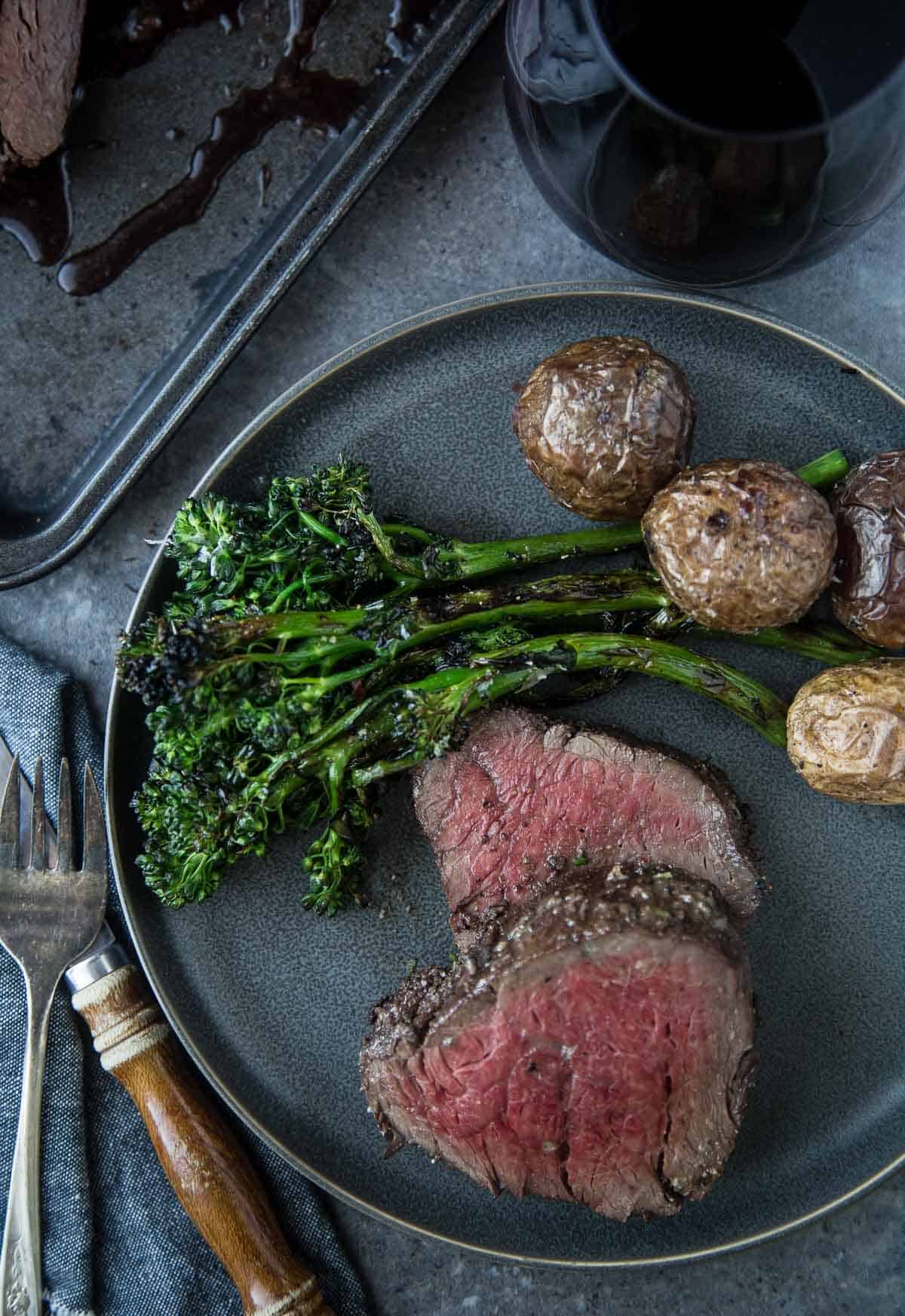 Slices of Smoked Beef Tenderloin on a plate with grilled broccolini and roasted potatoes and a glass of red wine