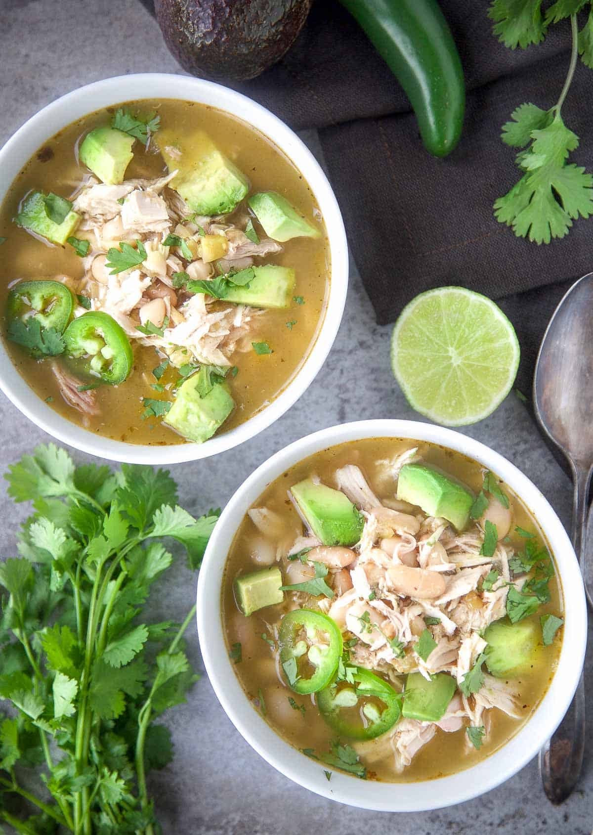 Two bowls of Smoked Salsa Verde Chicken Soup on a serving platter with wedges of lime and avocado