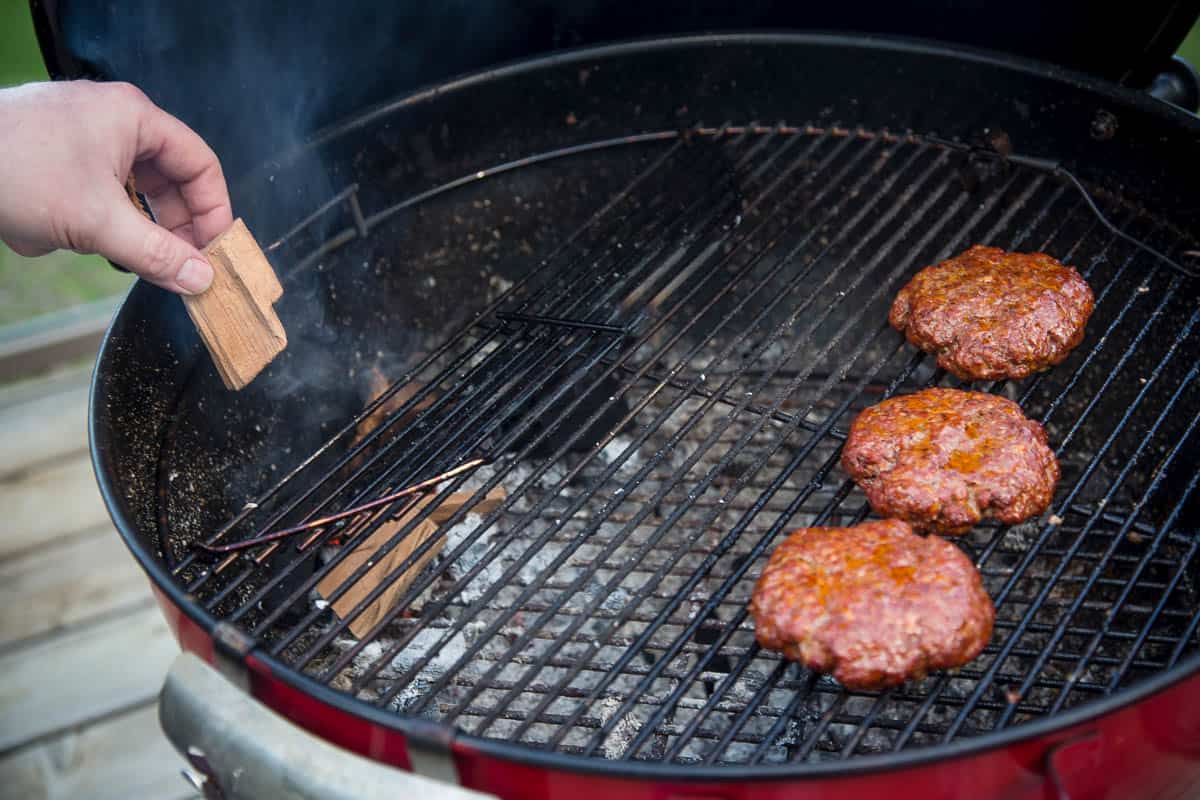 Adding wood chunks to a grill while three cheeseburgers cook over indirect heat