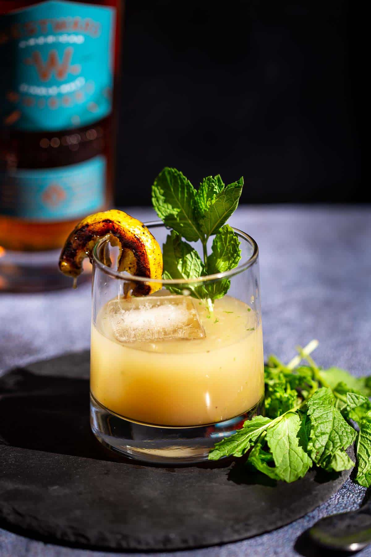 A Grilled Lemon Whiskey Smash in a glass and garnished with a grilled lemon wedge and fresh mint