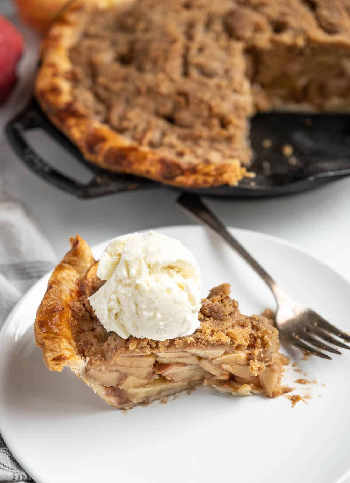 Slice of apple pie topped with ice cream with the pie in the background.