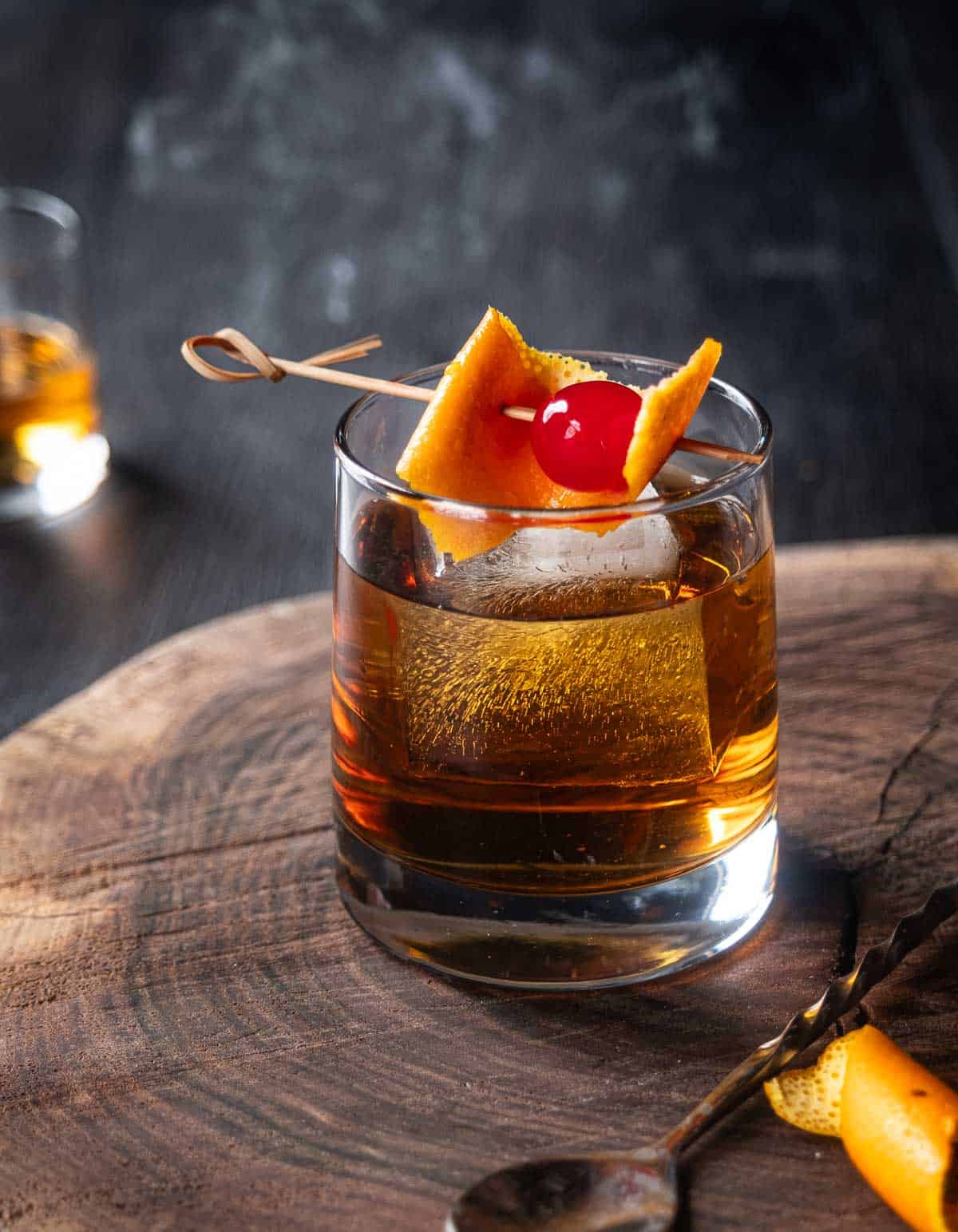 A smoked old fashioned cocktail garnished with a cherry and orange slice, and with a smoked ice cube