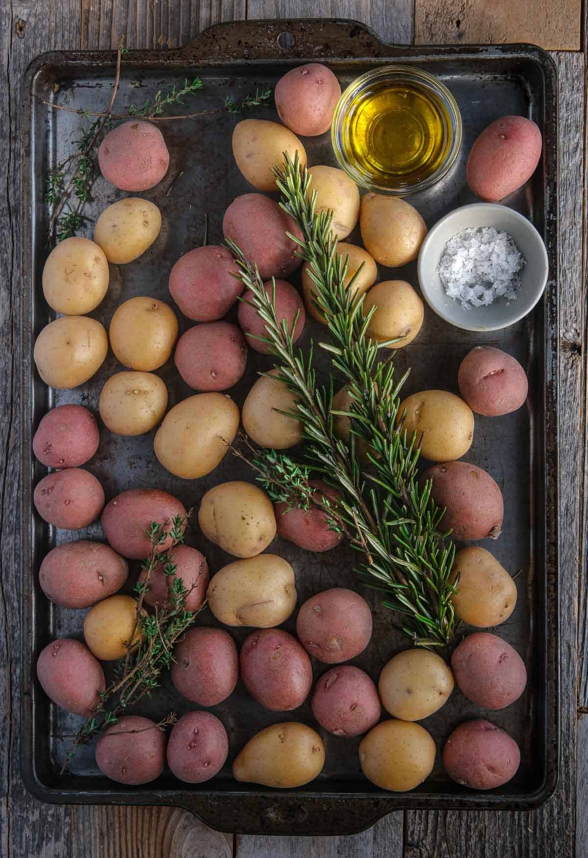 Ingredients for breakfast roasted potatoes on a sheet pan, including Yukon gold potatoes, olive oil, kosher salt, and fresh herbs