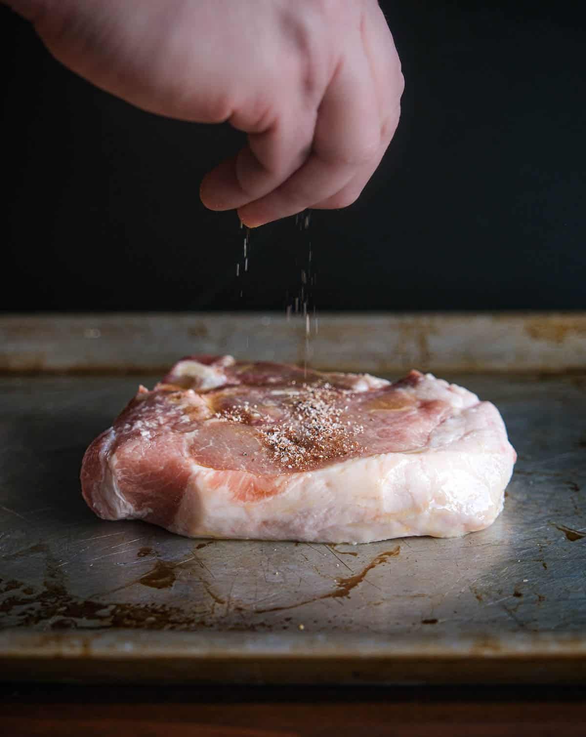 Seasoning a raw pork steak on a sheet pan. 