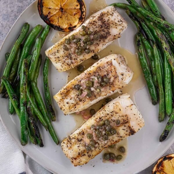3 Pieces of grilled halibut topped with a white wine sauce and surrounded by green beans on a plate.
