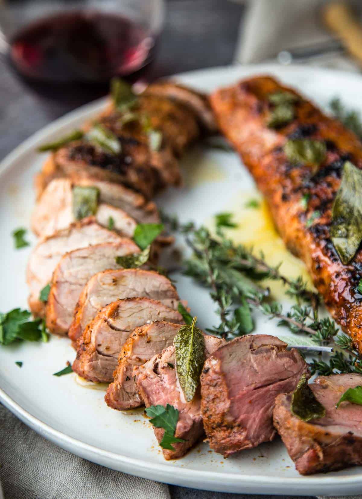 Slices of grilled pork tenderloin on a platter topped with sage butter sauce and a glass of wine.