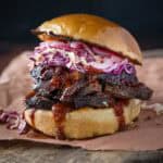 A smoked brisket sandwich on a cutting board with buns, BBQ sauce, coleslaw, and pickles.