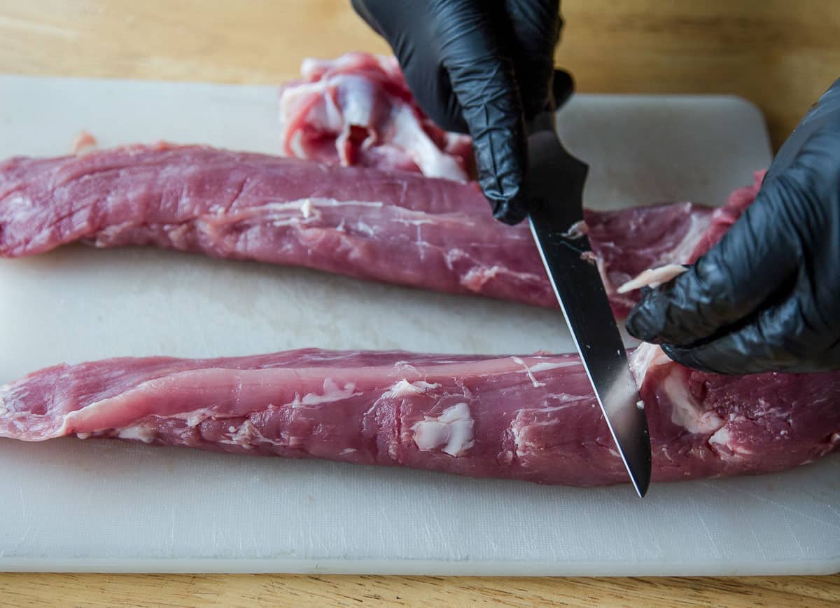 Trimming a pork tenderloin on a cutting board by removing the silver skin.