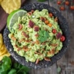 Smoked guacamole in a molcajete topped with bacon and cilantro surrounded by chips and greens.