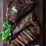 Two t bone steaks on a cutting board one sliced and topped with an herb compound butter.
