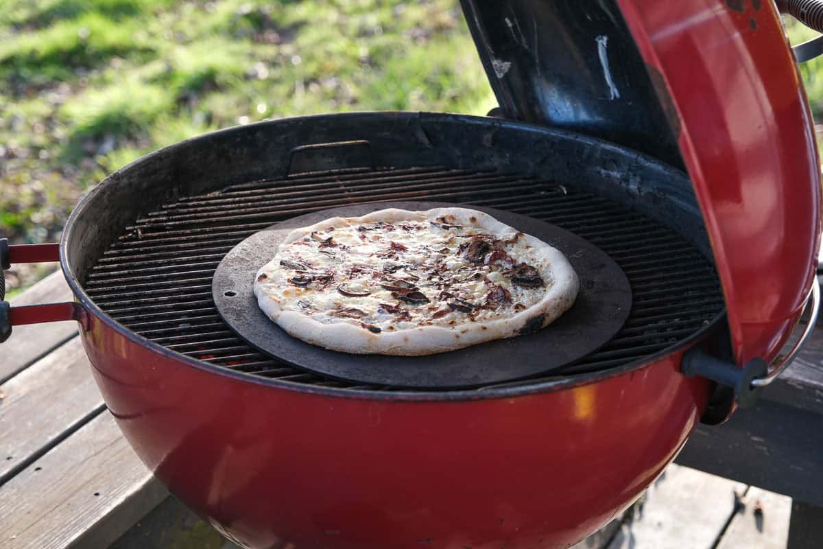 A pizza steel cooking a pizza on a weber kettle on the center of the grill grates.