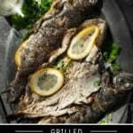 Three stuffed grilled trout on a plate with lemon and a side of white wine.