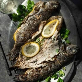 Three whole trout on a serving platter next to a glass of white wine.