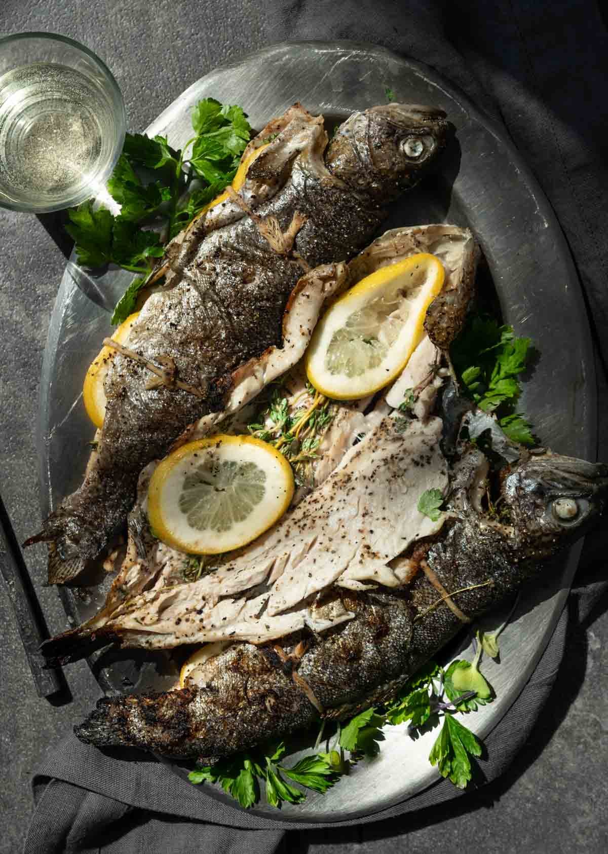 Three whole stuffed trout on a serving platter next to a glass of white wine.