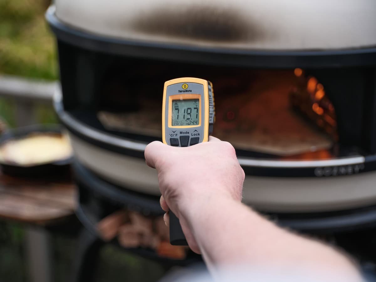 A hand holding an IR thermometer reading the temperature of a pizza stone in a pizza oven at 719 degrees Fahrenheit.