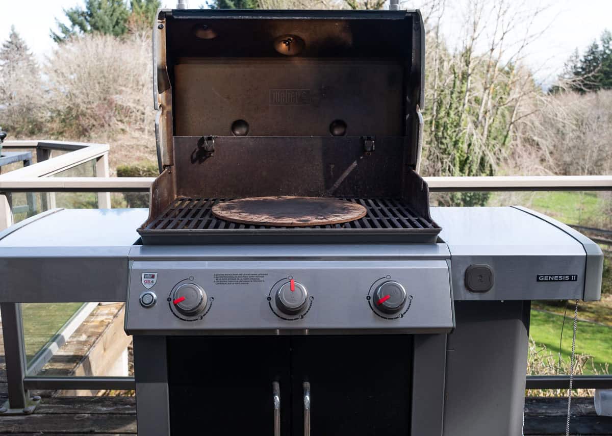 A pizza stone set in the middle grate of an open gas grill with no pizza on it.