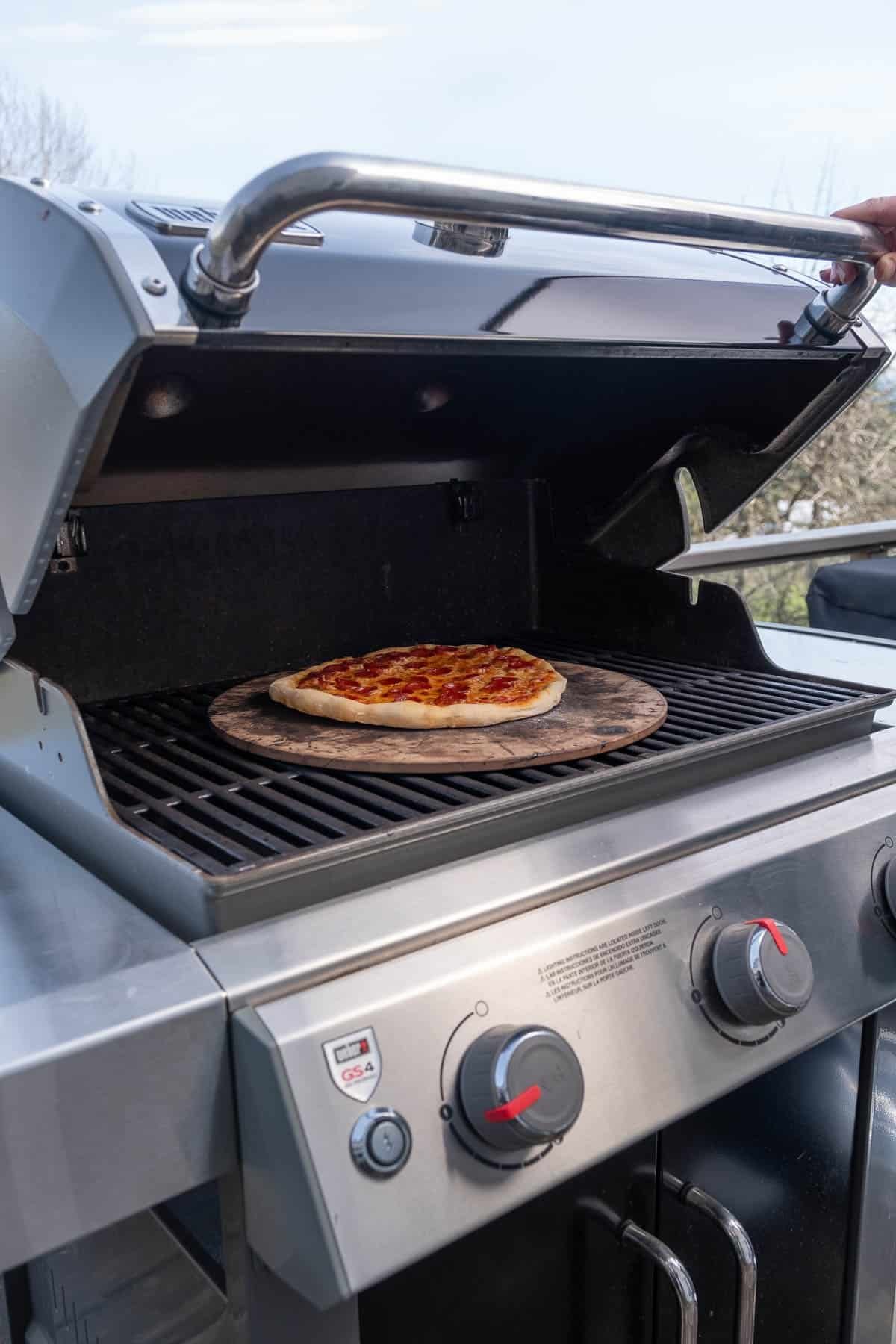 A pizza cooking on a pizza stone on a gas grill.