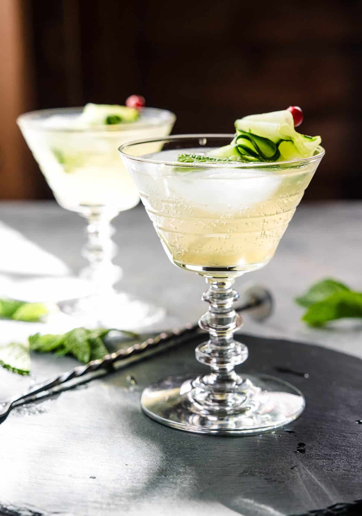 Two coupes filled with a cucumber spritz cocktail, topped with mint. and garnish.